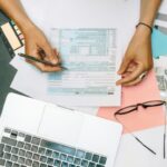 Hands writing on tax documents with laptop, glasses, and currency on desk.